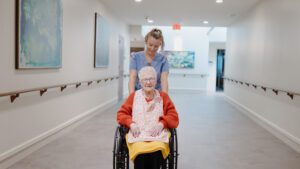 A woman being pushed down a hallway by a nurse at a skilled nursing facility